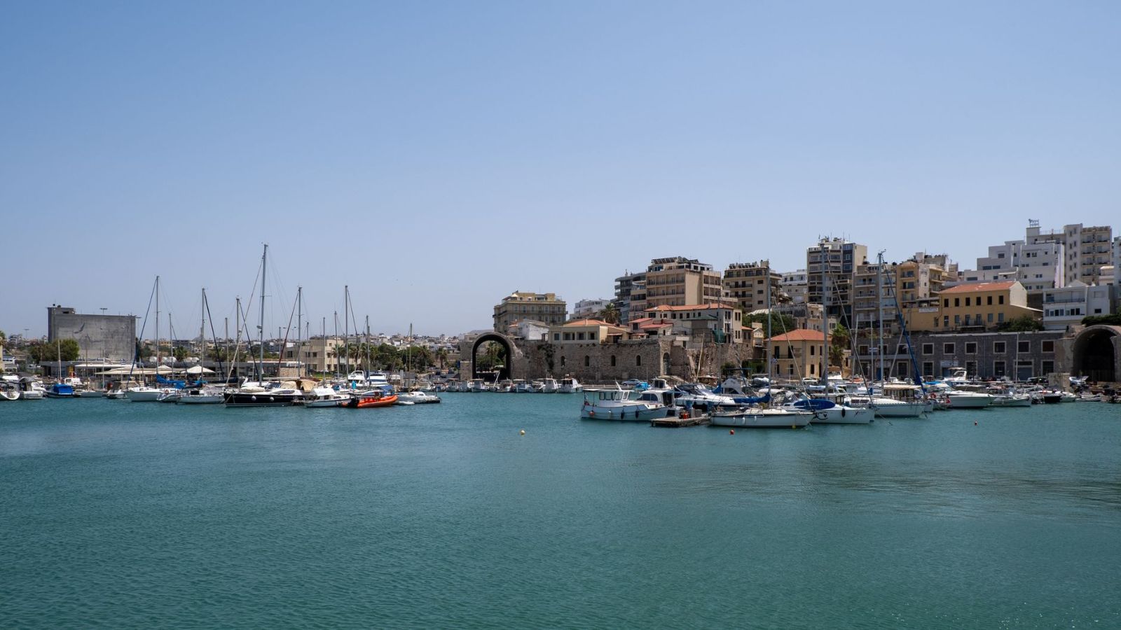 Beautiful view of Heraklion harbor with sailboats and cityscape under a blue sky.